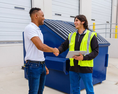 Business owner and waste service rep shaking hands near a commercial trash dumpster.